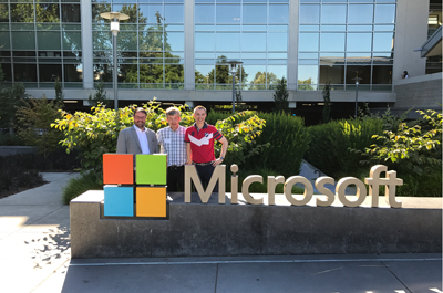 3 Personen stehend vor Haus mit Glasfront, hinter einer Skulptur Microsoft Logo