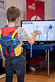 1 Kind mit Rücken zur Kamera, Blick Richtung Bildschirm, führt Übungen mit Hand aus