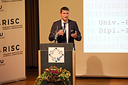 Mann stehend hinter Pult, Mikrofon in der Hand, Roll-up und Powerpoint-Präsentation im Hintergrund