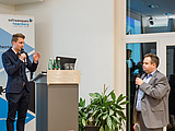 Person stehend hinter Rednerpult, hält Mikrofon in der Hand; daneben steht Moderator mit Mikrofon in der Hand